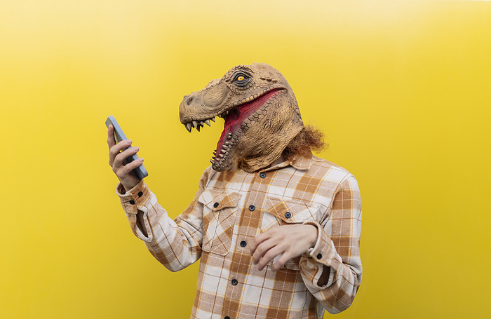 Man wearing dinosaur mask and using smart phone in front of yellow wall