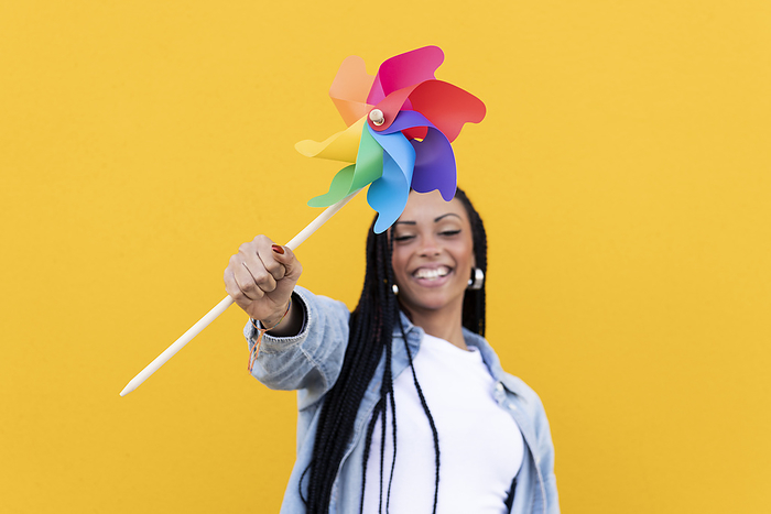 Happy young woman holding pinwheel toy in front of yellow wall