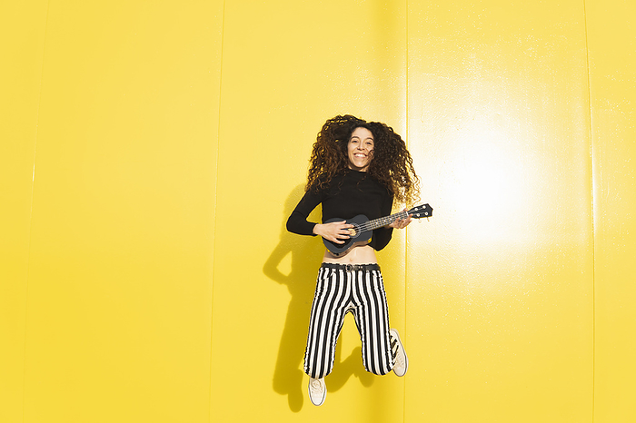 Smiling woman playing ukulele and jumping against yellow background