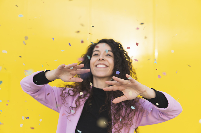 Smiling woman playing with confetti against yellow background