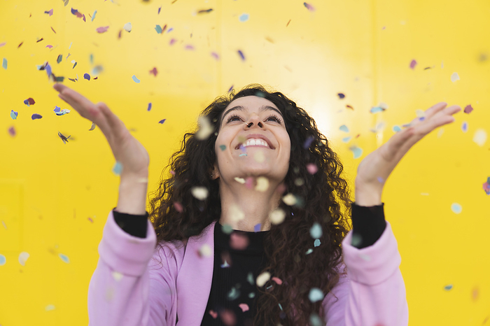 Cheerful woman playing with confetti against yellow background