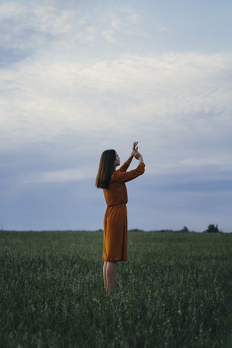 Woman with hands raised gesturing in field in front of sky at sunset