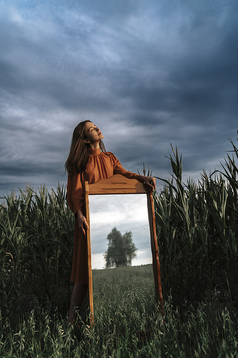 Woman standing with mirror in corn field at dusk