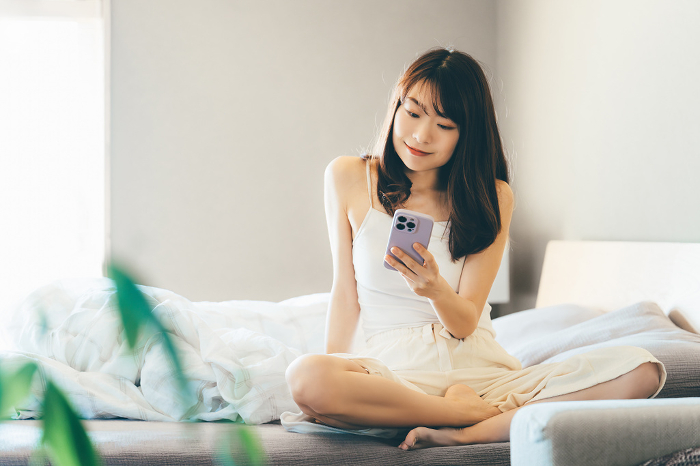 Smiling Japanese woman sitting on bed using smartphone (People)