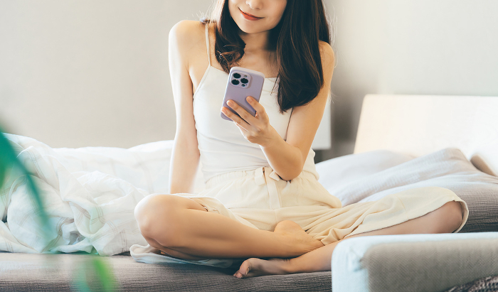 Smiling Japanese woman sitting on bed using smartphone (People)