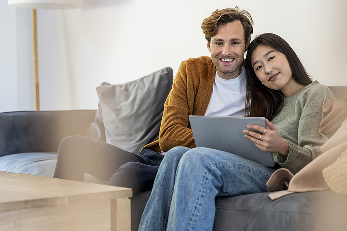Adult couple looking at the camera while sitting on sofa uding digital tablet