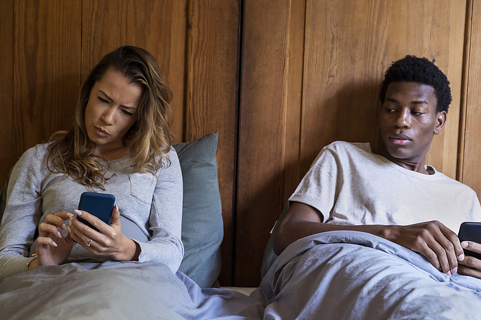 Distrustful boyfriend looking on girlfriend's smart phone while lying on bed