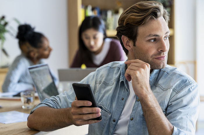 Young adult entrepreneur holding smart phone with coworkers on the background