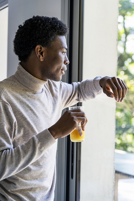 Side view of adult man leaning on window while drinking orange juice