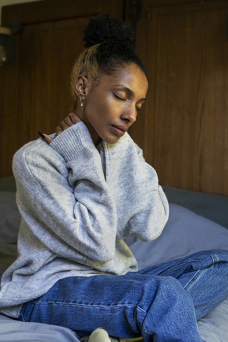 Stressed young adult woman touching her neck while sitting on bed