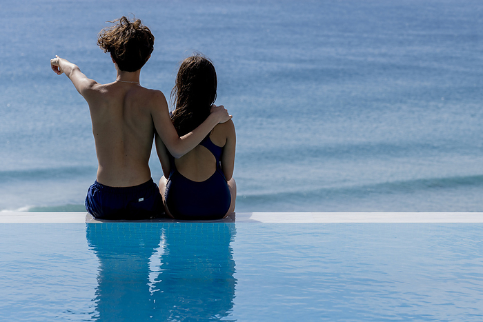 Rear view of young couple sitting at poolside and looking at ocean