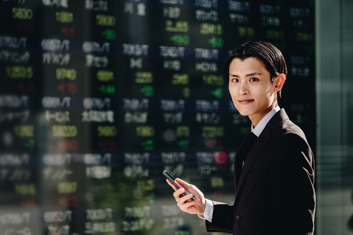 A Japanese man in his 30s looking at the camera as he trades stocks on NISA with his smartphone in front of the real-time stock price board on the electronic bulletin board (People)