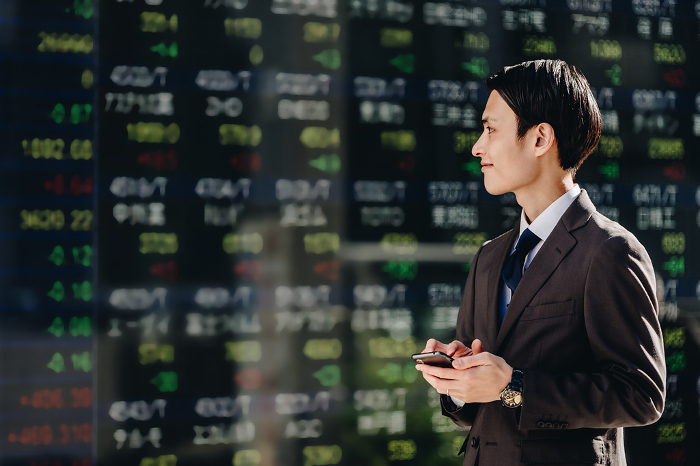 A Japanese man in his 30s trading stocks with a smartphone app in front of a real-time stock price board on an electronic bulletin board.