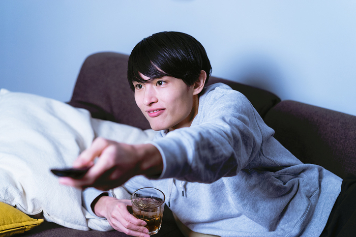 Lazy Japanese man in his 30s lying on the sofa, drinking and relaxing, operating the remote control and changing channels (People)