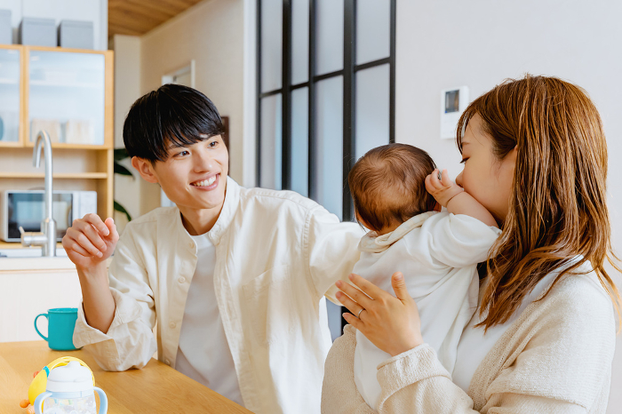 A mother woman holding her baby in her arms and a smiling father watching her in the living room of their home (Japanese family / People)