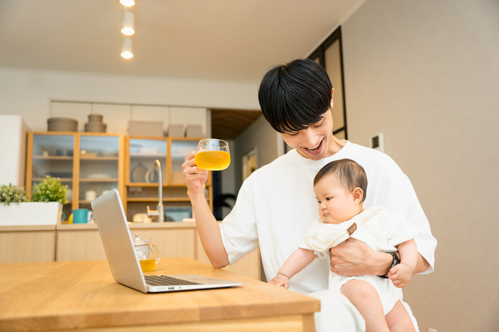 A Japanese man in his 20s or 30s working on his laptop while nursing his baby at his desk in the living room.