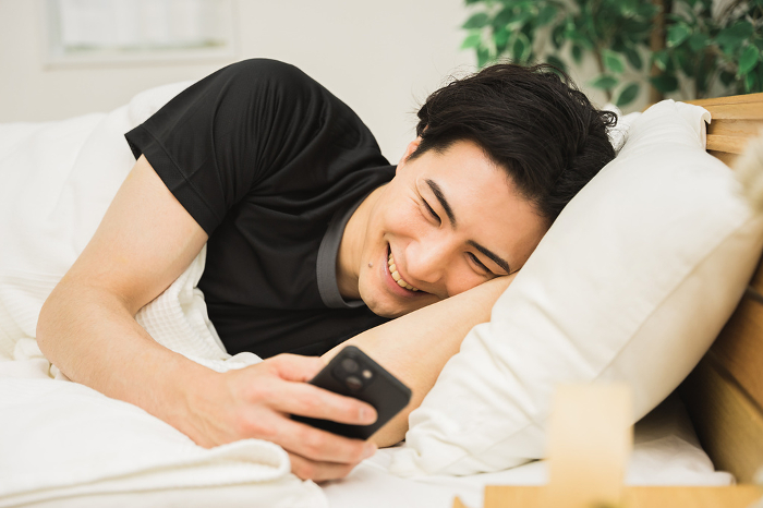 A Japanese man in his 20s or 30s operating a smartphone while lying on a bed in his room.