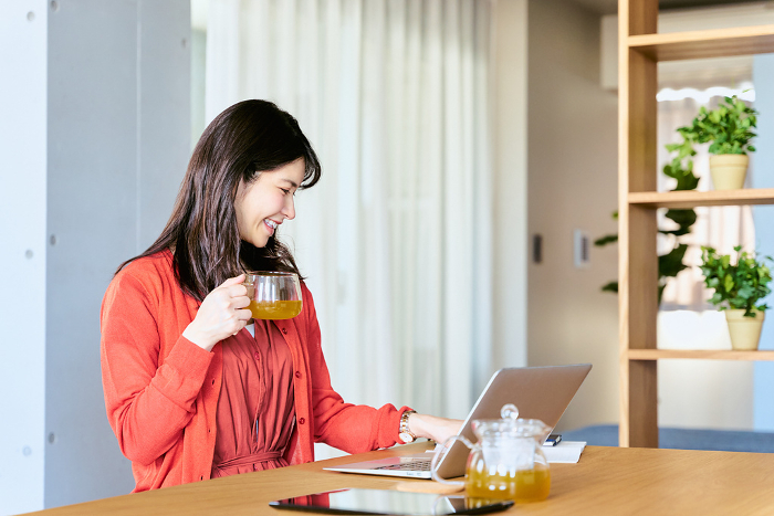 A Japanese woman in her 30s enjoys using her laptop in the living room to operate and research while drinking a cup of tea during a break from housework at home (People)