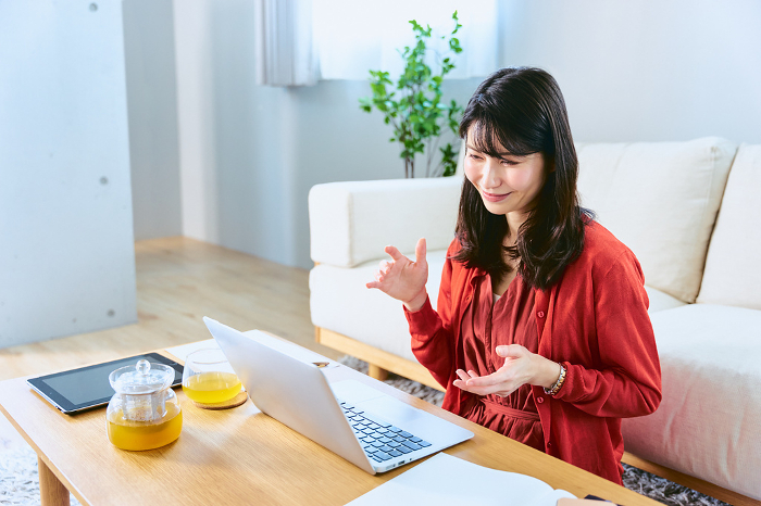 A Japanese woman in her thirties, sitting at a table in front of her living room sofa with her laptop open for online conversations and meetings (People)