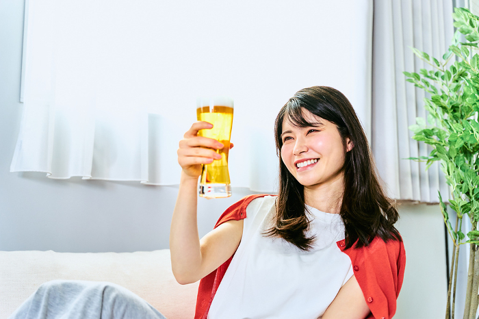 A Japanese woman with a fresh smile sitting on a sofa in her room at home in bright daylight, holding a beer in her hand and looking at it in the light.