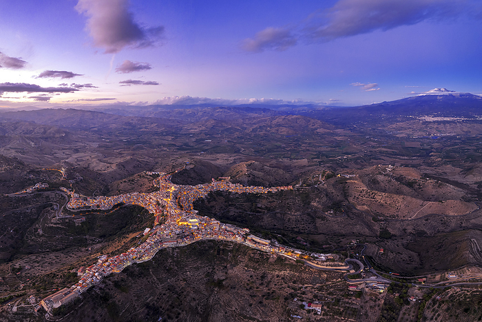 Panoramic aerial view of the man-shaped (or star-shaped) illuminated village of Centuripe with Etna volcano with snow in the background, dusk view, Centuripe, Enna province, Sicily, South of Italy, Italy, Europe