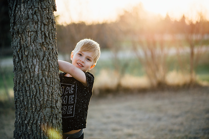 Close up of young boy smiling and peeking around tree in yard, by Cavan Images / Kimberli Fredericks