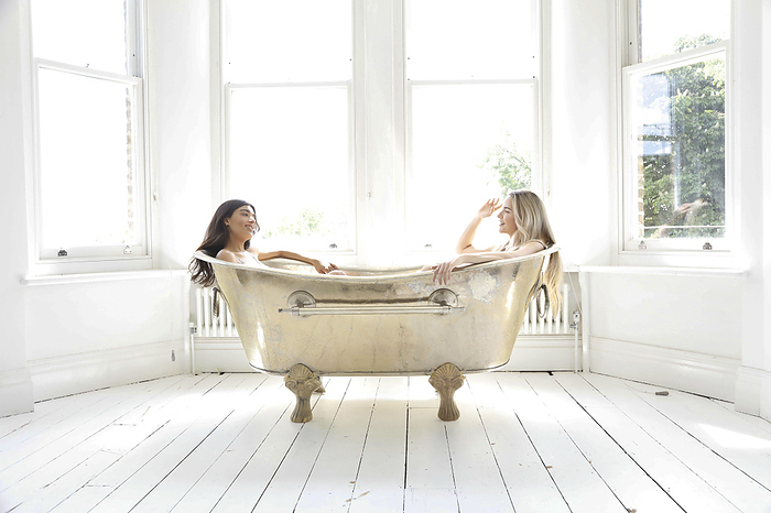 Two friends sharing a moment of laughter in a vintage clawfoot bathtub in a bright, sunlit room.