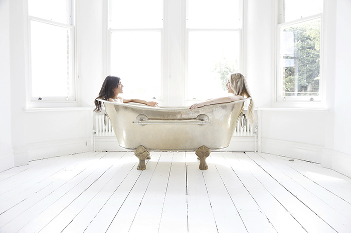 Two friends enjoying a relaxed chat in a vintage clawfoot bathtub in a bright sunlit room.