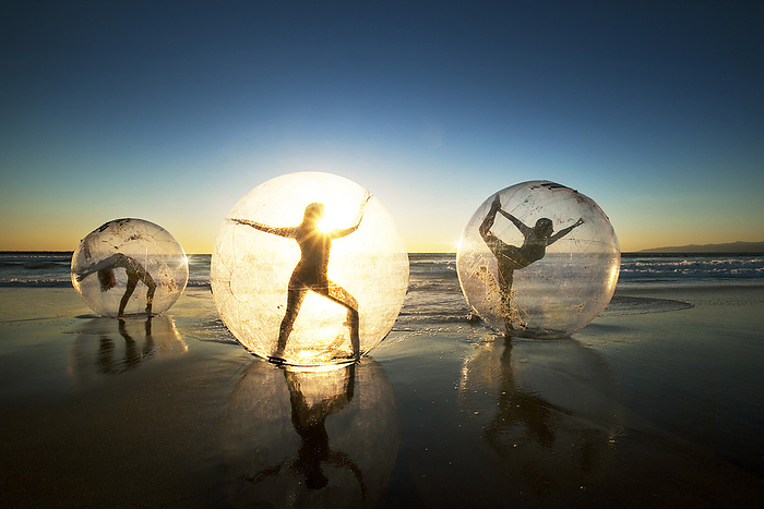 Silhouettes of people encased in transparent spheres on a beach at sunset.