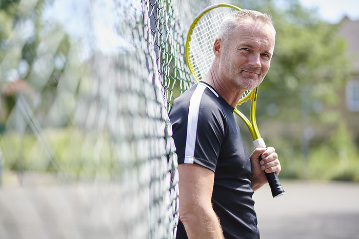 Mature man holding tennis racket leaning on fence
