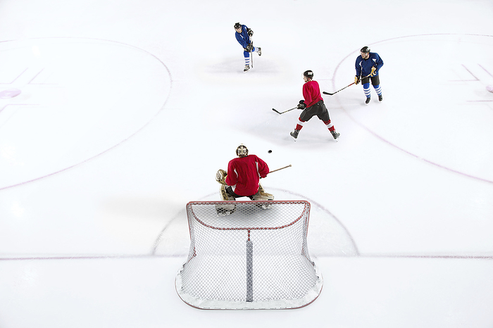 Hockey players in an intense game moment, with a focus on the goalie waiting for the incoming play.