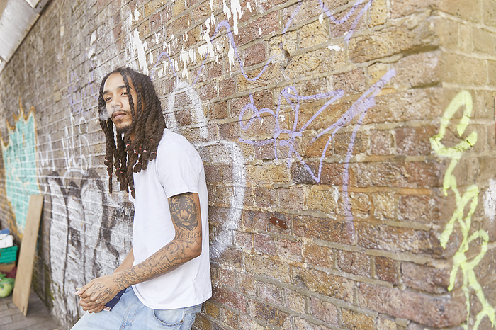 Young man with dreadlocks leaning against a graffiticovered brick wall