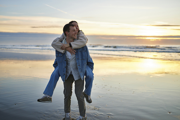Smiling boyfriend piggybacking girlfriend on beach at sunset