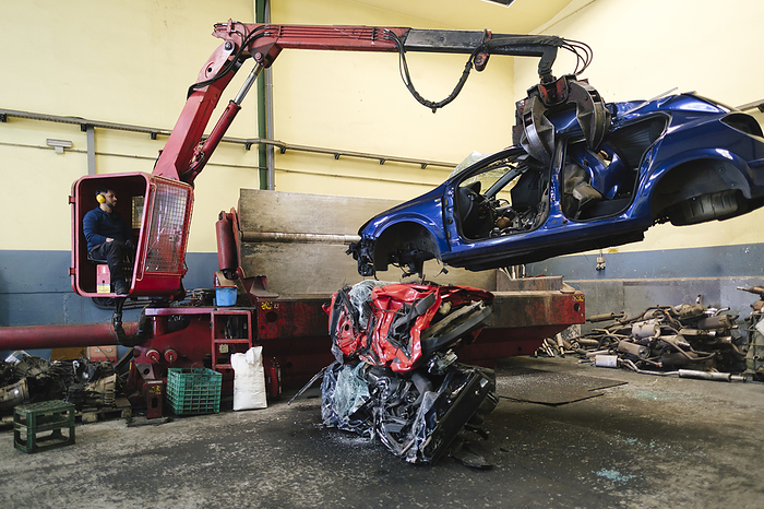 Mechanic operating crane machine and carrying scrapped car at workshop