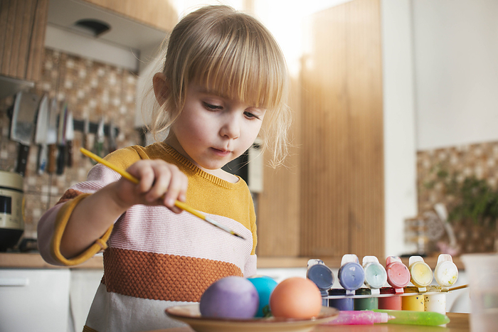 Cute girl coloring Easter eggs with paintbrush at home