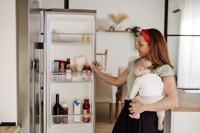 Smiling woman carrying baby daughter and opening refrigerator door