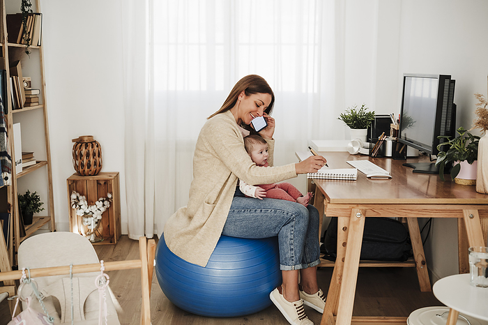 Mother sitting with baby girl and talking on mobile phone in home office