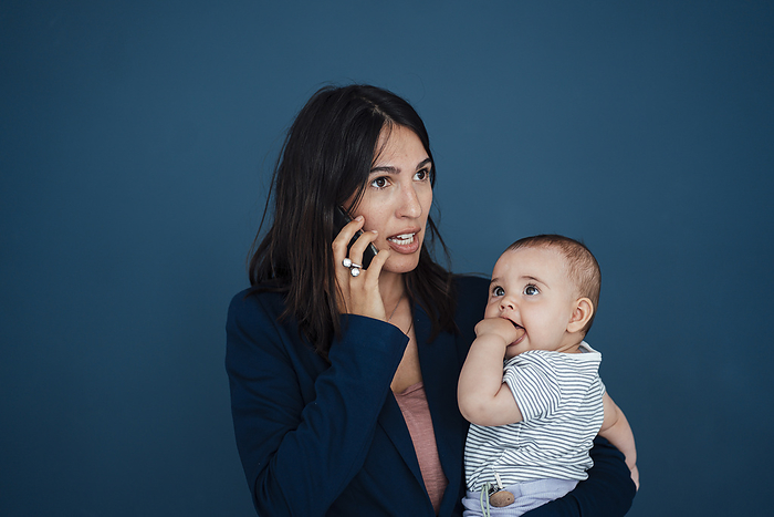 Busy businesswoman carrying daughter talking on mobile phone in front of blue background