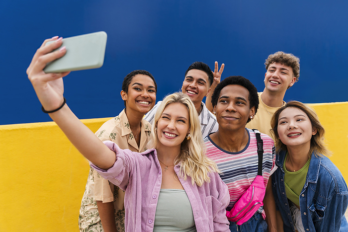 Laughing multi-ethnic friends taking smartphone selfie in front of yellow wall