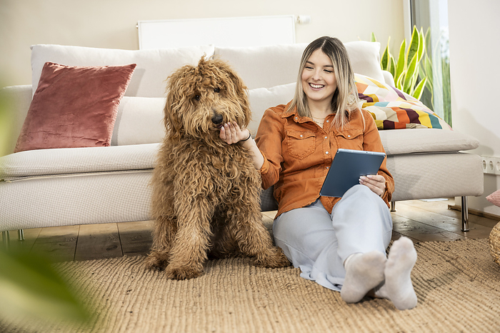 Happy beautiful woman holding tablet PC and stroking dog at home