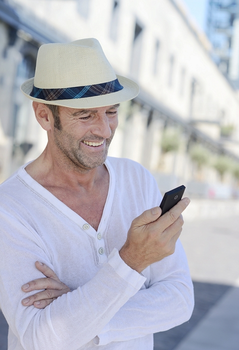 Portrait of smiling mature man with stubble and summer hat using smartphone