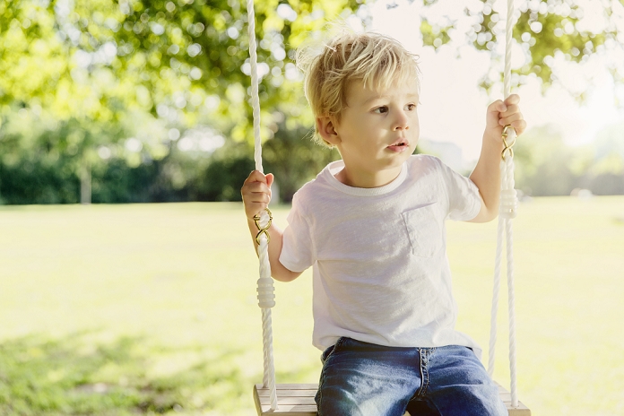 Germany, Bonn, Male toddler sitting on swing under tree in nature, looking away