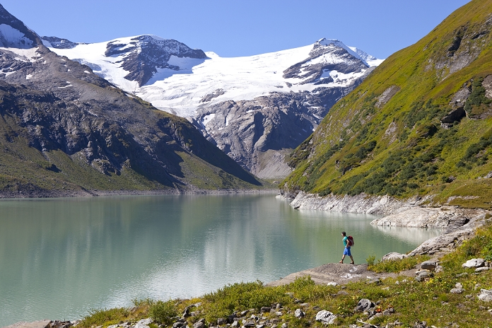 A male hiker near the Mooserboden lake, during the Glocknerrunde, a 7 stage trekking from Kaprun to Kals around the Grossglockner, the highest mountain of Austria. A glacier of the Grossglockner range is in the background and melting water feeds the lake, providing Hydroelectricity.    Located in the heart of the Hohe Tauern National Park which contrasts impressive, glacier-covered high alpine scenery with an alpine farming landscape carefully cultivated for centuries, the Glockner Circuit offers a unique nature and walking experience.    At the end of each day?s walk, mountain shelters or alpine inns beckon to rest, eat and get a good night?s sleep. Alpine huts are equipped and operated only with the basic essentials because of the difficulties in bringing in supplies and disposing of waste, the altitude, the extreme climate and the often long ascents! After any stage of the walk, you have the opportunity to leave the Glockner Circuit and walk down to the National Park villages of Kaprun, Uttendorf, Kals, Heiligenblut or Fusch to spend the night in a guesthouse, inn or hotel.     The tour mainly spans high alpine terrain but can be negotiated by anyone with some alpine experience and corresponding fitness and equipment, as there are no glaciers or roping sections along the circuit. A head for heights and surefootedness are necessary. Photo by Menno Boermans / Aurora Photos