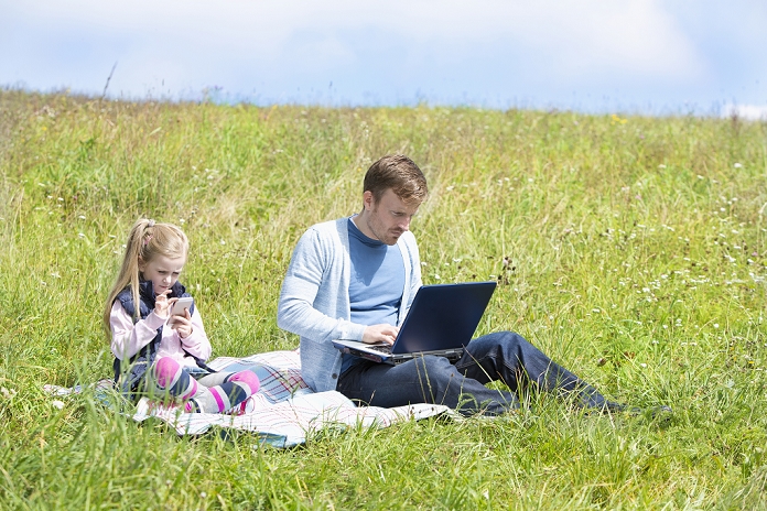 Father and daughter sitting on meadow, smartphone and laptop