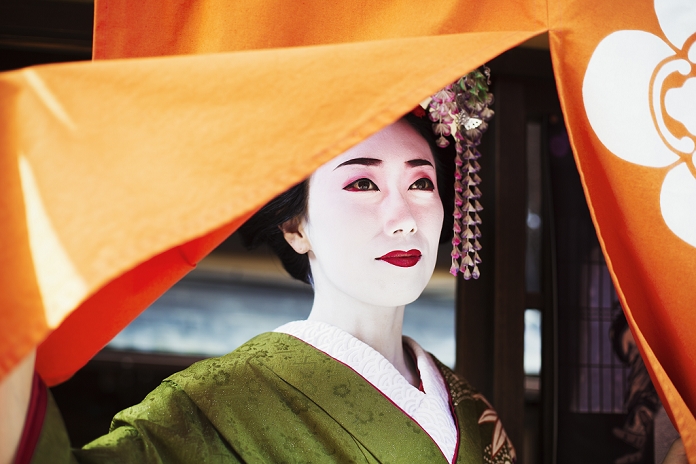 apprentice geisha A woman dressed in the traditional geisha style, wearing a kimono with an elaborate hairstyle and floral hair clips, with white face makeup with bright red lips and dark eyes lifting an orange curtain.