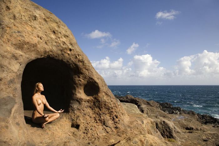 Young nude Asian woman mediatating in cave with coastline of Maui, Hawaii.