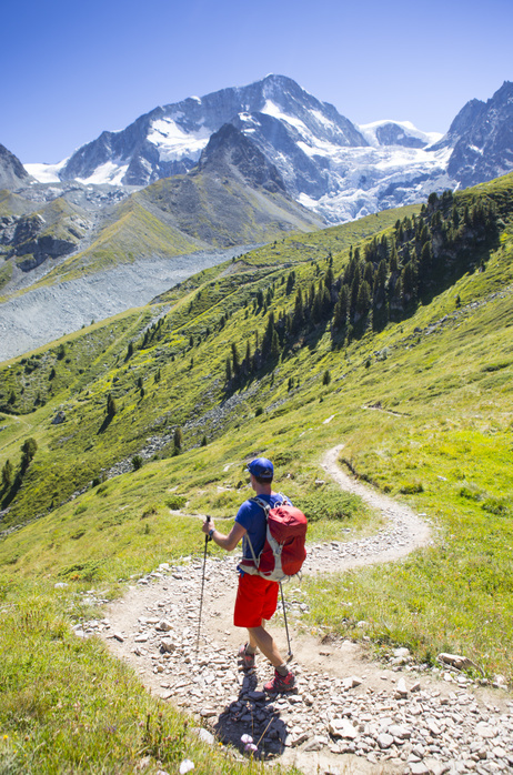 A colorful dressed male hiker is descending curved mountain trail in a green summer landscape with glaciers in the background.  A colorful dressed male hiker is descending curved mountain trail in a green summer landscape with glaciers in the background.  