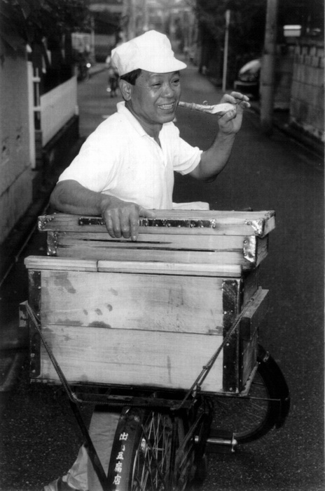 Tofu shop  Date of photo unknown   20th Century Graffiti  A Bugle Calling Sunset   Tokyo, Izukawa san making his rounds in an alley. When called, he stops his bicycle and cuts tofu with a knife.