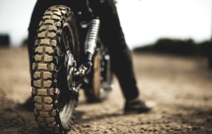 Rear view of man sitting on cafe racer motorcycle on a dusty dirt road, close up of tire. Rear view of man sitting on cafe racer motorcycle on a dusty dirt road, close up of tire.