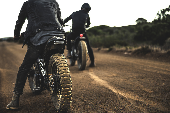 Rear view of two men sitting on cafe racer motorcycles on a dusty dirt road. Rear view of two men sitting on cafe racer motorcycles on a dusty dirt road.